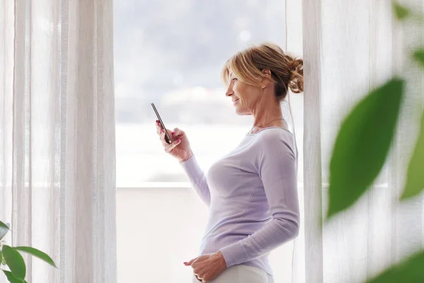 Woman Standing Next Window Looking Away She Holding Her Smartphone — ストック写真