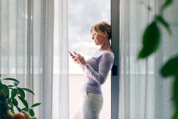 Happy Mature Woman Standing Next Window Home Connecting Her Smartphone — ストック写真