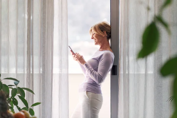 Happy Mature Woman Standing Next Window Home Connecting Her Smartphone — ストック写真
