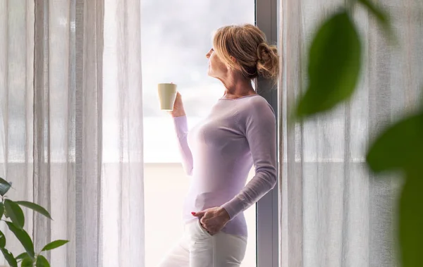 Mature Woman Relaxing Home Standing Next Window She Looking Away — Stok fotoğraf