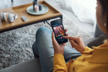 Woman sitting on the couch at home and streaming podcasts on her smartphone