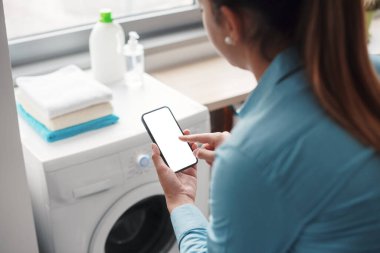 Woman using smart appliances at home, she is connecting the washing machine to her phone, blank display on the smartphone
