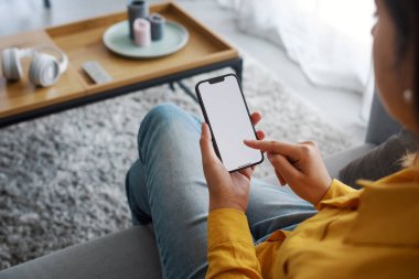 Woman sitting on the couch at home and using her smartphone, blank display