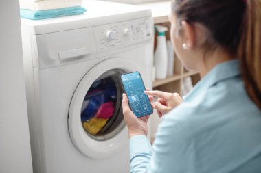 Woman using a smart washing machine, she is controlling the appliance from her smartphone