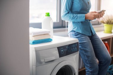 Woman doing laundry at home using a smart washing machine, she is controlling the smart washer from her smartphone