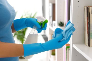 Woman cleaning a bookcase at home, she is spraying detergent on a cloth, hands close up