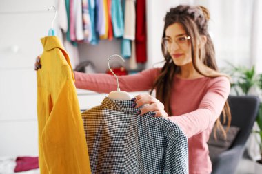 Young happy woman holding two shirts and comparing them, she is choosing what to wear