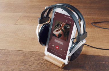 A pair of headphones and a digital tablet on a wooden table. On the display you see the player, and a music album playing. Concept of new ways to relax anywhere.
