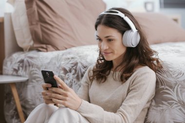 Young woman relaxing on the bed, she is wearing headphones and watching videos on her smartphone
