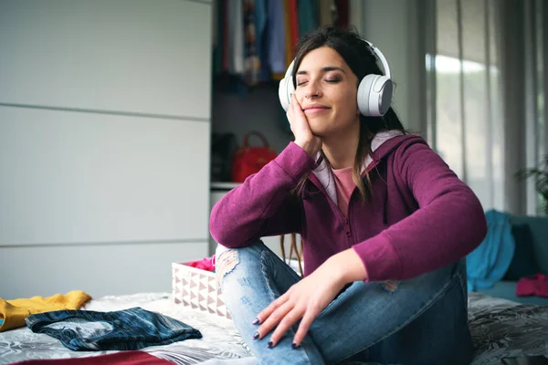 Happy relaxed woman sitting on the bed and listening to music, she is wearing headphones