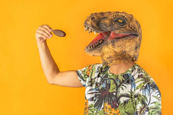 Man in dinosaur animal head mask eating chocolate ice cream isolated on yellow background with copy space