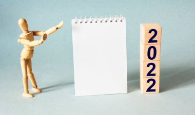 Business goal with year numbers and target icon sign on a stack of wooden cube blocks on a blue background with a notepad for writing and a wooden puppet.