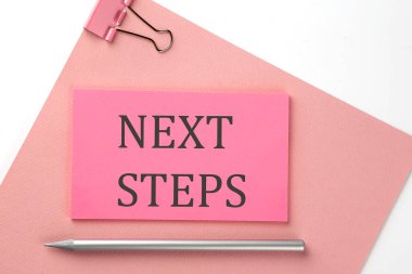 Pembe kağıt üzerine pembe etiket, beyaz zemin üzerinde kalem ve metin NEXT STEPS