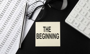 Concept THE BEGINNING message on sticker with glasses,chart and calculator on black background