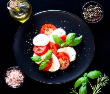 Dilimlenmiş domatesli İtalyan caprese salatası, mozzarella peyniri, fesleğen, siyah tabakta zeytinyağı koyu ahşap arka planda baharatlar.