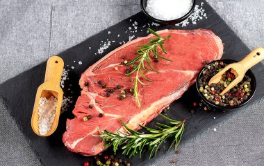 Kara tahtada çiğ biftek, biberiye, biber ve ahşap kaşıkta tuz. Gri arkaplan.