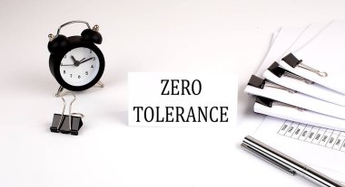 Üzerinde ZERO TOLERANCE yazan kart beyaz arka planda, ofis malzemeleri ve çalar saatin yanında. İş dünyası