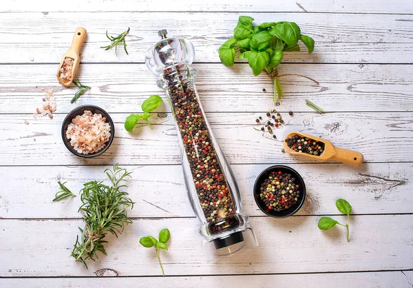 Pepper mill with different peppers in bowl with himalayan salt and fresh rosemary and basil on wooden background