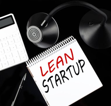 Siyah arkaplan üzerinde kalem, hesap makinesi ve masa lambası olan Lean STARTUP metni ile açılan kağıt defter.