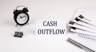 Beyaz arka planda CASH OUTFLOW yazılı kart, ofis malzemeleri ve çalar saatin yanında. İş konsepti.