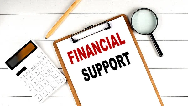 FINANCIAL SUPPORT words on clipboard, with calculator, magnifier and pencil on white wooden background