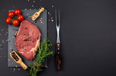 Çiğ et, siyah arka planda biftek, ahşap kaşıkta baharatlar, biberiye ve çatal, üst manzara.