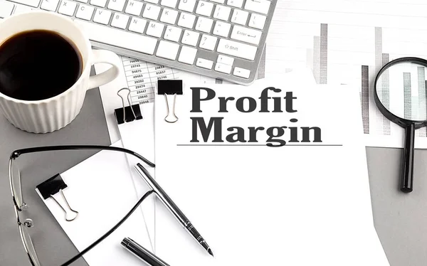 PROFIT MARGIN text on paper with magnifier, coffee and keyboard on grey background