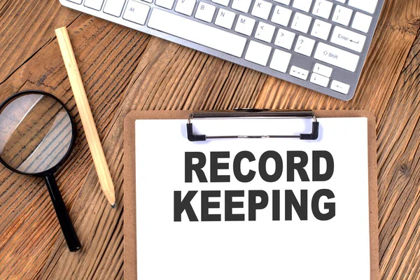 RECORD KEEPING text on paper clipboard with magnifier and keyboard on a wooden background