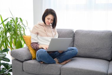 Gülümseyen genç kadın dizüstü bilgisayar kullanıyor, evdeki kanepede oturuyor.