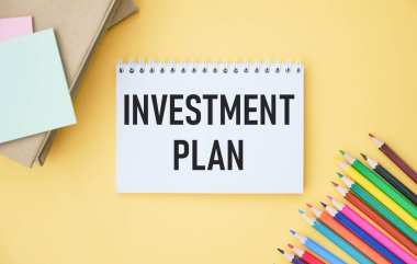 Hafif bir arkaplanda, beyaz bir çalar saat, bir hesap makinesi, bir kalem ve INVESTMENT PLAN yazılı bir defter. Düz yatıyordu