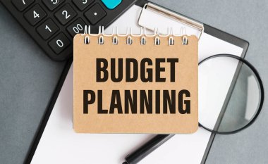 Beyaz kağıt üzerinde bütçe planlama metni. Defterdeki yazıyı. İş konsepti. Gri bir arka planda not defteri. gözlük, kalem, kalem, saat, hesap makinesi