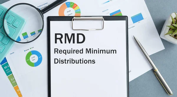 Masada raporlar, bir büyüteç, bir hesap makinesi, bir kalem ve içinde RMD gerektiren Minimum Dağıtımlar olan beyaz bir defter var.