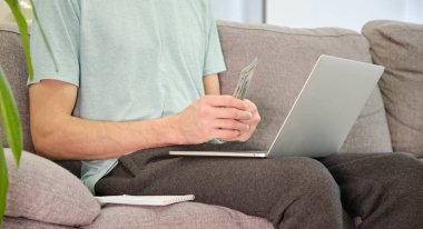 Genç adam dizüstü bilgisayarla koltukta oturuyor ve faturalarını hesaplayan kağıtlarla.