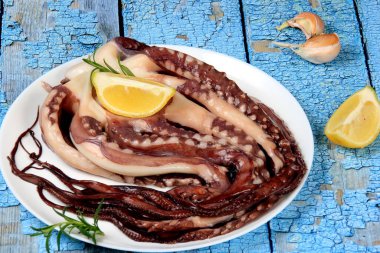 Limon, yeşil limon, biberiye ve marul yapraklarıyla süslenmiş beyaz bir tabakta çiğ ahtapot. Mavi arka plan. Üst görünüm.