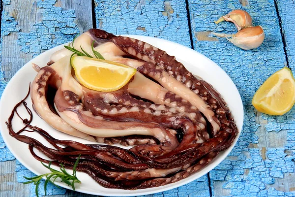 Limon, yeşil limon, biberiye ve marul yapraklarıyla süslenmiş beyaz bir tabakta çiğ ahtapot. Mavi arka plan. Üst görünüm.