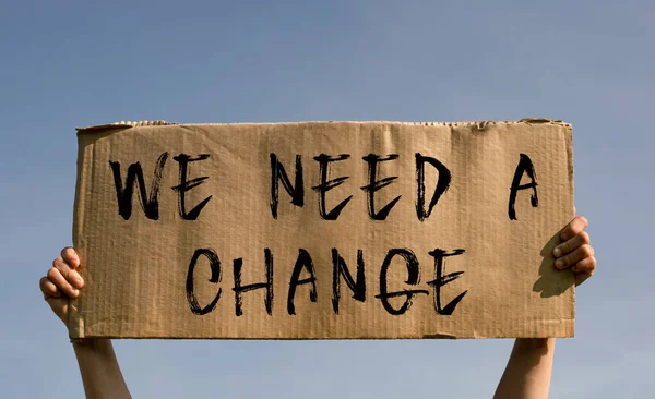 A poster with the words WE NEED A CHANGE in the hands of a young girl against a blue sky.