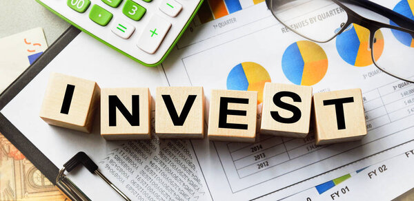 Надпись на кубиках, слово INVEST на кубиках деревянной текстуры. Вид сверху. Рабочий стол с документами. Надпись на финансовой, деловой или экономической теме.