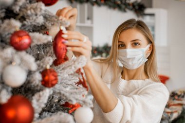 Sarışın genç bir kadın Covid-19 için tıbbi maske takarak Noel ağacını süslüyor. Coronavirus ve Noel konsepti, güzel tatil geçmişi