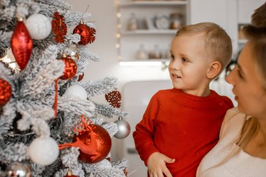Beyaz süveter içinde güzel dekore edilmiş Noel ağacının yanında küçük oğlunu tutan çekici bir anne.