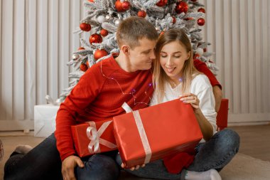 Noel ağacının yanında hediye kutularıyla oturan mutlu genç çift. Genç adam kadınını kucaklıyor ve kadın hafifçe dilini gösteriyor..