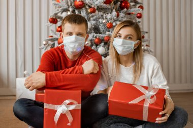 Koruyucu maskeli kadın ve erkek Noel ağacının yanında oturmuş hediyelerini tutuyorlar ve kameraya bakıyorlar. Covid-19 salgını sırasında uzak bir Yeni Yıl kutlaması