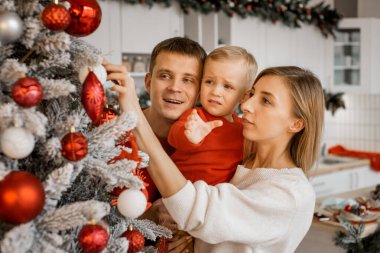 Mutlu anne, baba ve küçük oğlu evde birlikte Noel ağacı süslüyorlar, küçük çocuk ağlıyor ve oyuncak topu koluna almak istiyor.