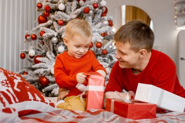 Baba ve küçük oğlu, Noel ağacının yakınındaki evde, rahat yatak odasında kırmızı süveter içinde hediyelerini açıyor..