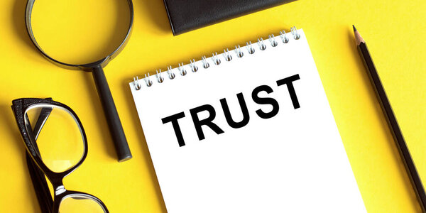 TRUST text on notepad with pencil, glasses and magnifier on yellow table