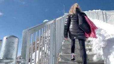 Ceketli, güneş gözlüklü ve maskeli yetişkin bir kadın merdivene tırmanıyor, arkasını dönüyor ve Glacier 3000 'de ellerini neşeyle açıyor. Turizm ve seyahat kavramı.