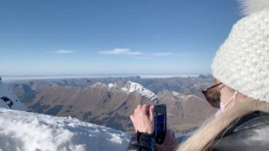 Yünlü şapkalı ve güneş gözlüklü sarışın kadın Glacier 3000 'de akıllı telefonuyla fotoğraf çekiyor. Kış yolculuğu konsepti