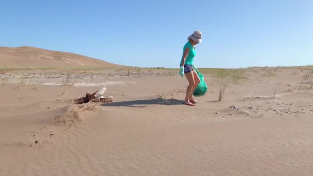 Beyaz şapkalı ve yeşil eldivenli olgun bir kadın yeşil bir çantada kum tepeciklerinden plastik çöp topluyor.. — Stok video