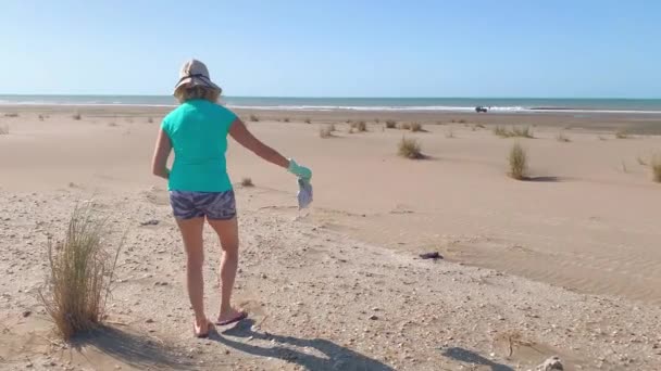 Reife Frau mit einem grünen Müllsack, der im Sand in der Nähe des Meeres liegengelassenen Abfall aufhebt. Pflege des Erdkonzepts. — Stockvideo