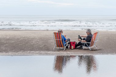 İki olgun gri saçlı adam plaj sandalyelerinde oturmuş bir eşe su döküyorlar. Arka planda denizin dalgaları.