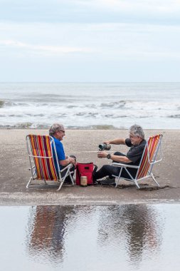 İki olgun gri saçlı adam plaj sandalyelerinde oturmuş arkadaşa su döküyorlar, gülümsüyorlar ve kameraya bakıyorlar..
